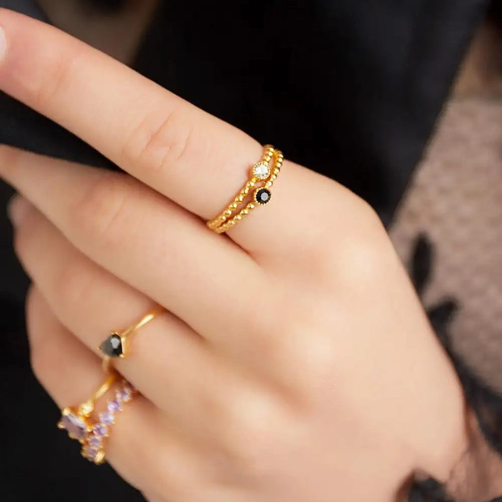 A close-up of a hand wearing multiple gold rings, including a textured band with a small black stone and a delicate ring with a black oval stone.