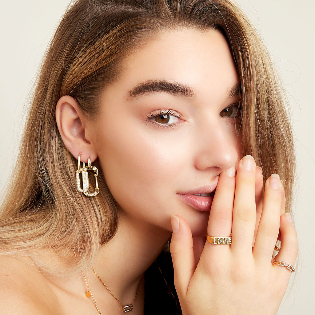 A close-up lifestyle shot of a woman with blonde hair wearing gold jewelry, including a gold hoop earring and gold rings with 'LOVE' lettering.