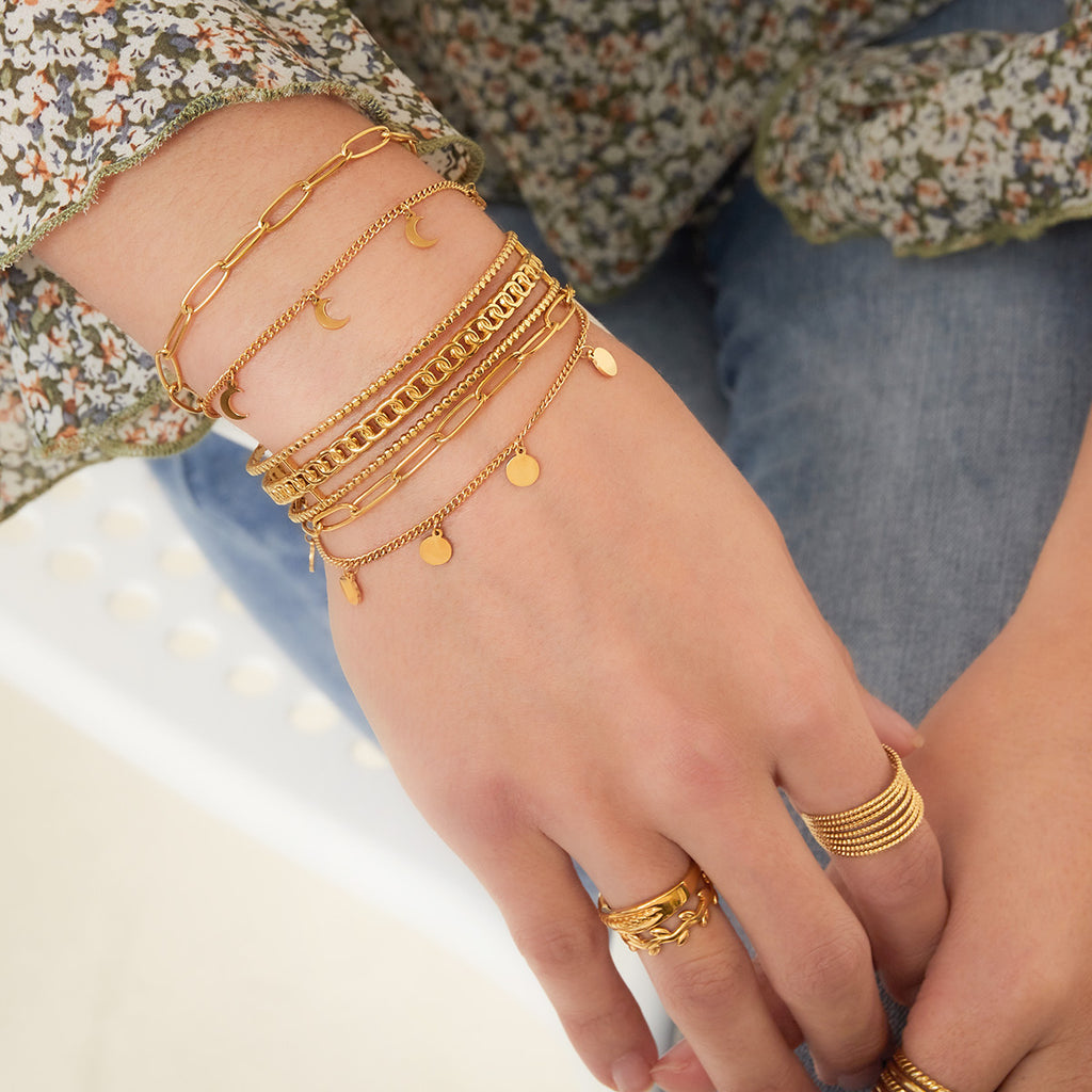 A close-up of a person's hand and wrist wearing multiple gold bracelets, including chain styles with crescent moon and disc charms, along with gold rings, against a soft floral top and blue jeans background.