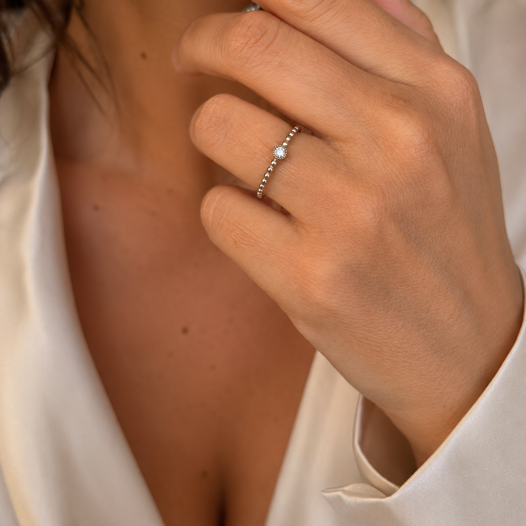 A close-up of a delicate silver ring with a single round clear gemstone worn on a person's finger, set against a soft cream-colored satin top.