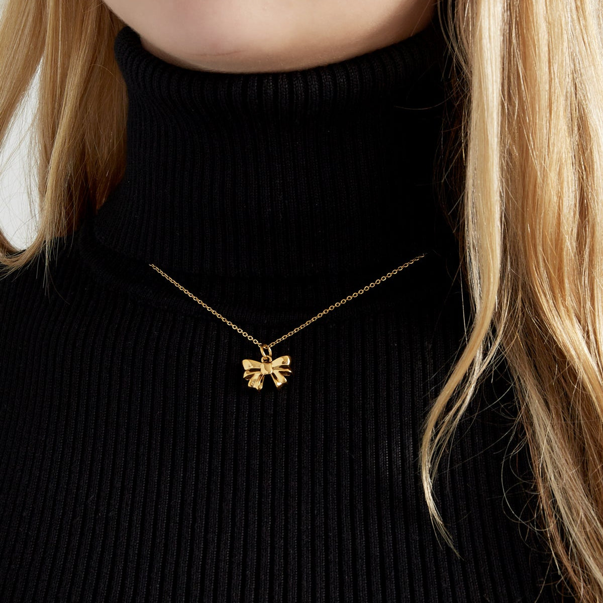 Gold necklace with a bow pendant worn by a person in a black turtleneck.