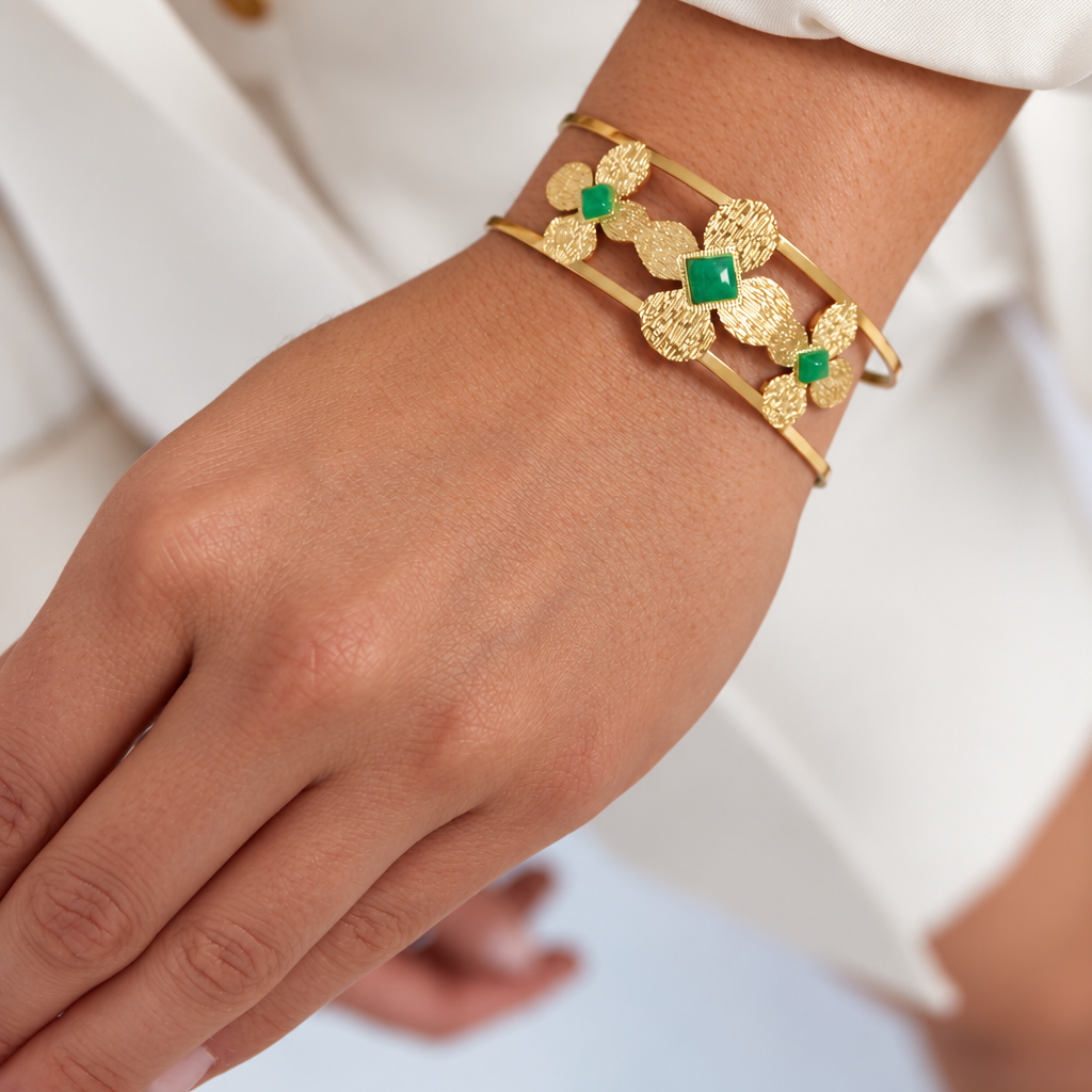 A close-up of a person's wrist wearing a gold cuff bracelet with textured floral motifs and small green square accents, paired with a white blazer.